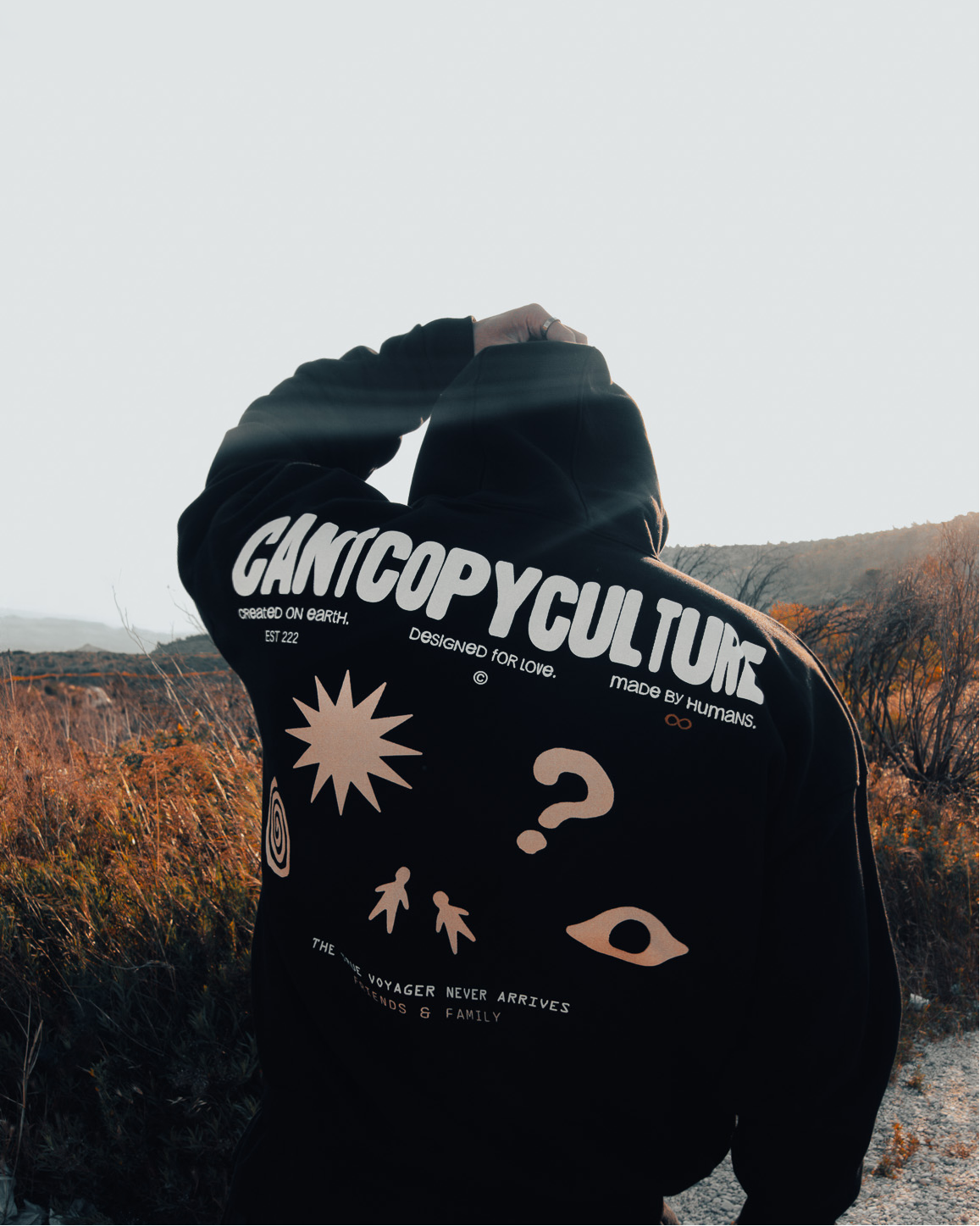 A person standing outdoors on a gravel path wearing a black hoodie with a large back graphic that reads 'GAMICOPYCULTURE' and includes white and peach illustrations, with hills and dry brush in the background.